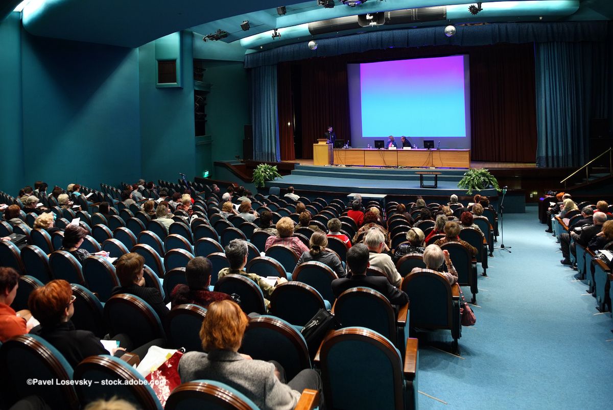 A group of people in an auditorium watch two experts give a presentation. Image credit: ©Pavel Losevsky – stock.adobe.com