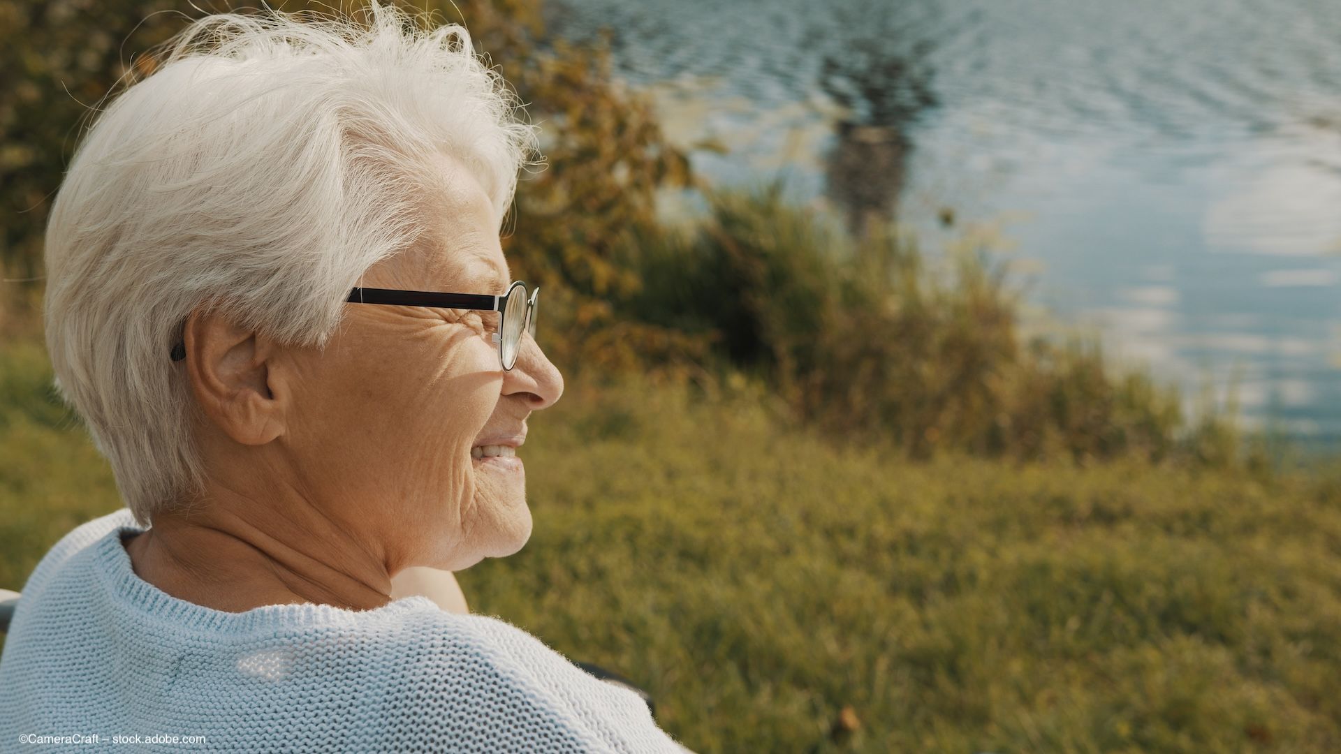 An elderly woman with glasses sits on a grassy hillside, near a tranquil blue lake. Concept image for geographic atrophy and vision-related quality of life measures. ©CameraCraft – stock.adobe.com