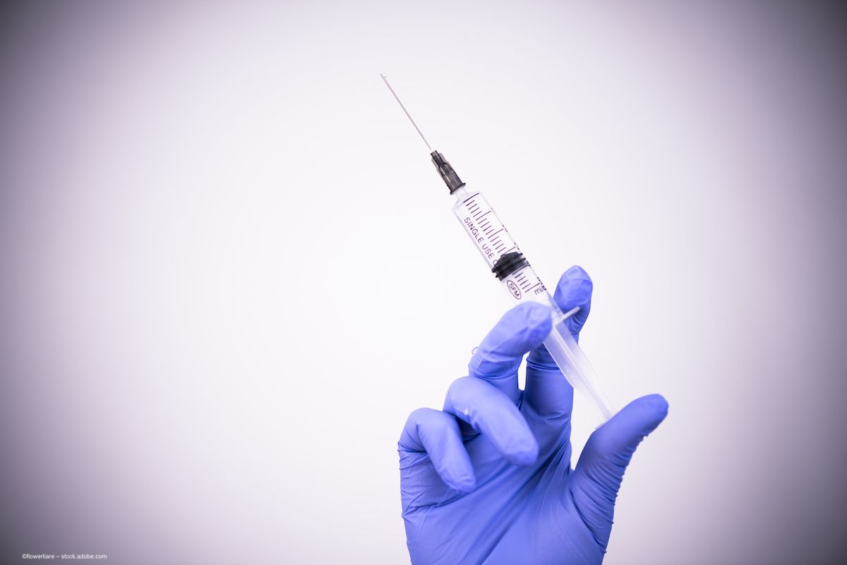 A hand wearing latex surgical gloves holds a syringe against a stark background. Image credit: ©flowertiare – stock.adobe.com