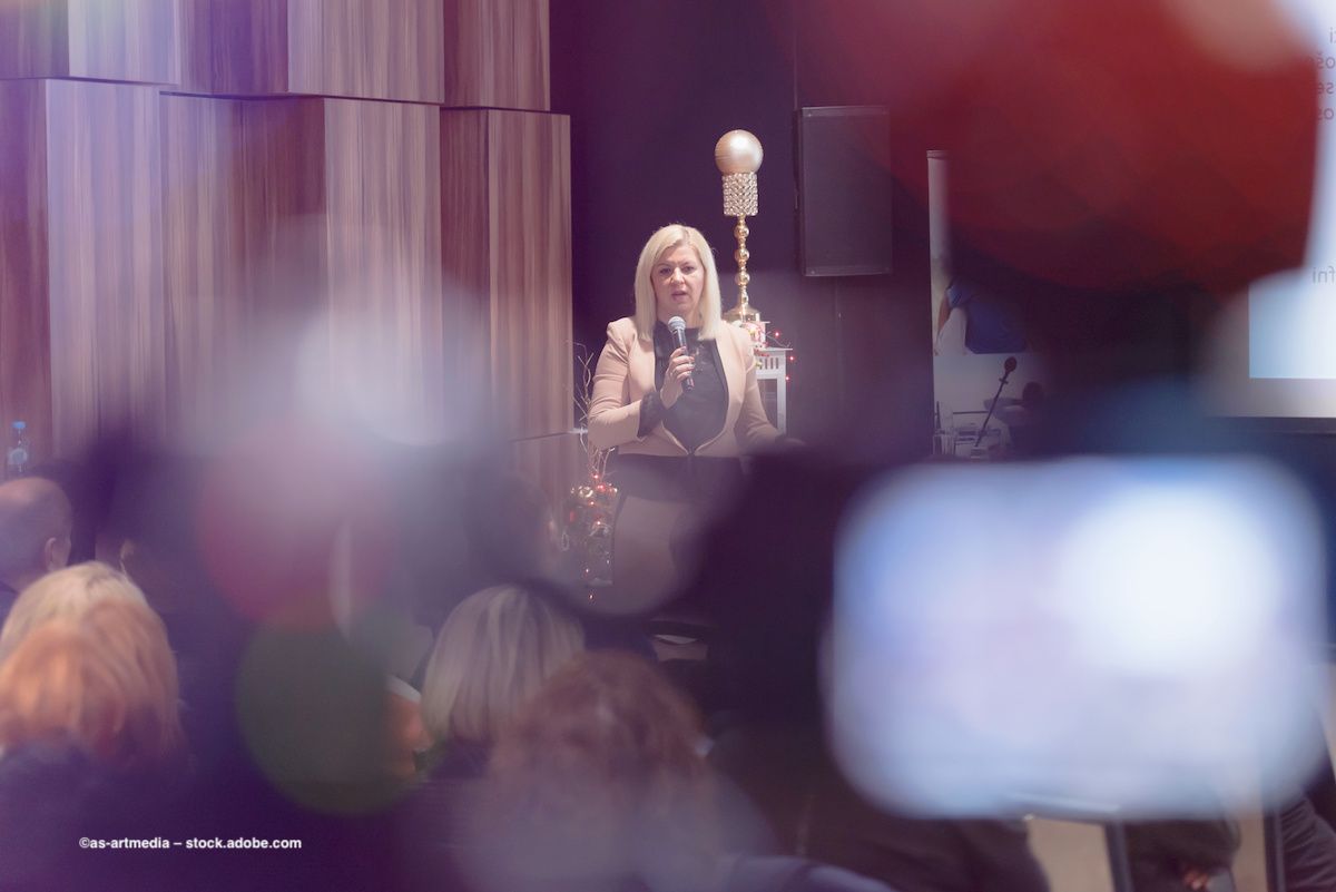 A woman stands at a lectern delivering a presentation to an audience. She is viewed through the blur of the crowded audience. Image credit: ©as-artmedia – stock.adobe.com
