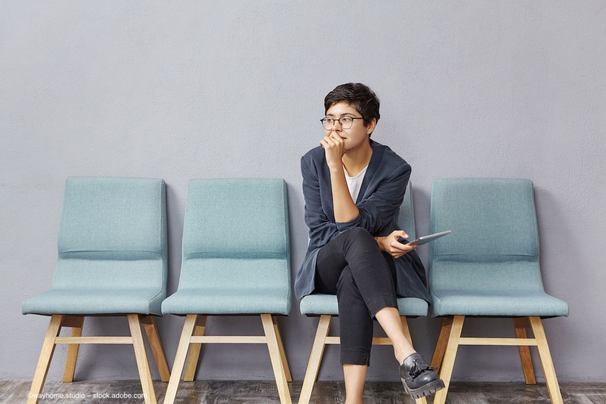 A young person with short androgynous hair sits in a waiting room, looking thoughtful. Concept image for racial groups in clinical trials and representation in studies. Image credit: ©wayhome.studio – stock.adobe.com