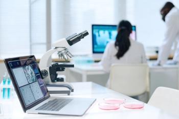 A microscope, petri dishes and a laptop sit in a laboratory, with researchers in the near distance. Image credit: ©pressmaster – stock.adobe.com