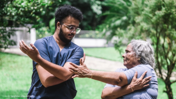 A nurse in scrubs and glasses helps his elderly patient stretch and exercise. Image credit: ©Jacob Varghese – baseimage