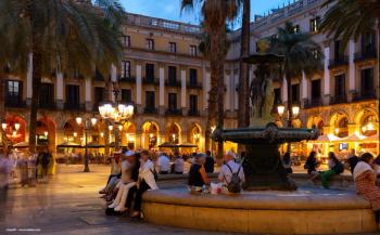 Placa Reial in Barcelona, Spain. A fountain is lit up by streetlamps. Tourists gather at night. Image credit: ©JackF – stock.adobe.com