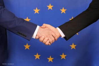 Two business people in suits shake hands in front of a European Union flag. Concept image for Optegra and EssilorLuxottica acquisition, European expansion, expand into new areas of medical technology. Image credit: ©New Africa – stock.adobe.com