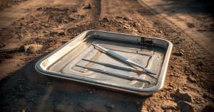 A surgical tray sits on a dirt road. There are visible scalpels and other metal tools. Concept image for ocular trauma, conflict and combat zones, eye surgery and emergency ophthalmology in low-resource, high-conflict regions or war zones. Created by Jennifer Toomey using Adobe Firefly AI.