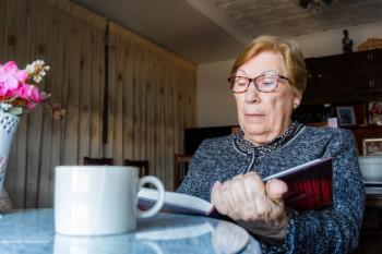 An older woman with glasses on sits and reads a book, a mug of coffee in front of her. Concept image for geographic atrophy lesions (GA) and age-related macular degeneration (AMD). Image credit: ©MiguelAngel – stock.adobe.com