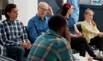 A group of patients, including a diverse array of men, women and children, sit in a waiting room. Image credit: ©DC Studio – stock.adobe.com