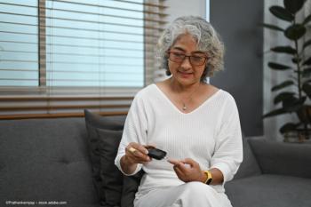 An older woman sitting on a sofa measures her blood glucose by using a finger-prick blood test. Image credit: ©Prathankarnpap – stock.adobe.com