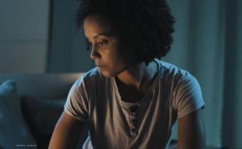 A woman sits in a dark room, looking sadly at the floor. Image credit: ©StockPhotoPro – stock.adobe.com
