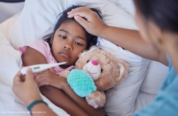 A sick child in bed holds her stuffed teddy bear. An adult takes the sick child's temperature on a digital thermometer. Image credit: ©Alexis Scholtz/peopleimages.com – stock.adobe.com