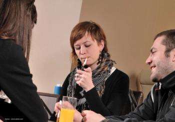 A small group of people sit at a table. One person lights a cigarette. Image credit: ©sima – stock.adobe.com