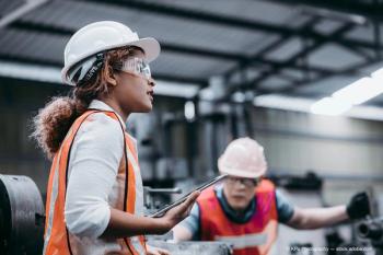 Female industrial engineer wearing a white helmet while standing in a heavy industrial factory behind she talking with workers, Various metal parts of the project (Adobe Stock / KPs Photography)