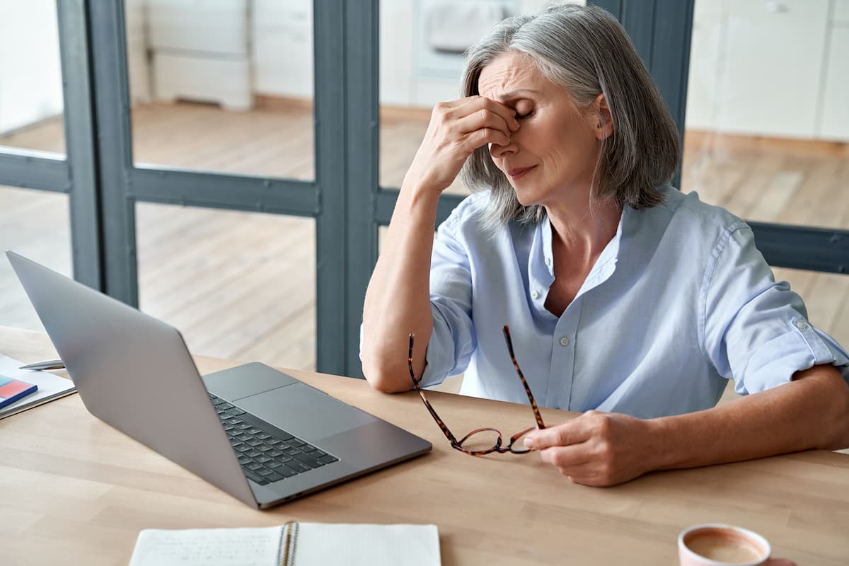 Older woman holding bridge of nose at desk Image credit: AdobeStock/insta_photos