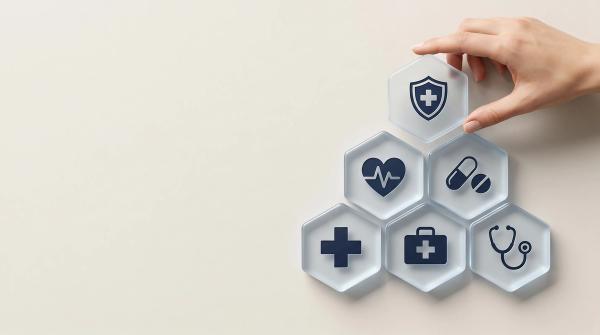 Hand placing a hexagonal tile with a shield and cross icon on a pyramid of health and medical service symbols, representing comprehensive healthcare and insurance. | © Abimusa Gallery - stock.adobe.com (2073964658)
