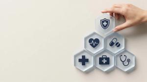 Hand placing a hexagonal tile with a shield and cross icon on a pyramid of health and medical service symbols, representing comprehensive healthcare and insurance. | © Abimusa Gallery - stock.adobe.com (2073964658)