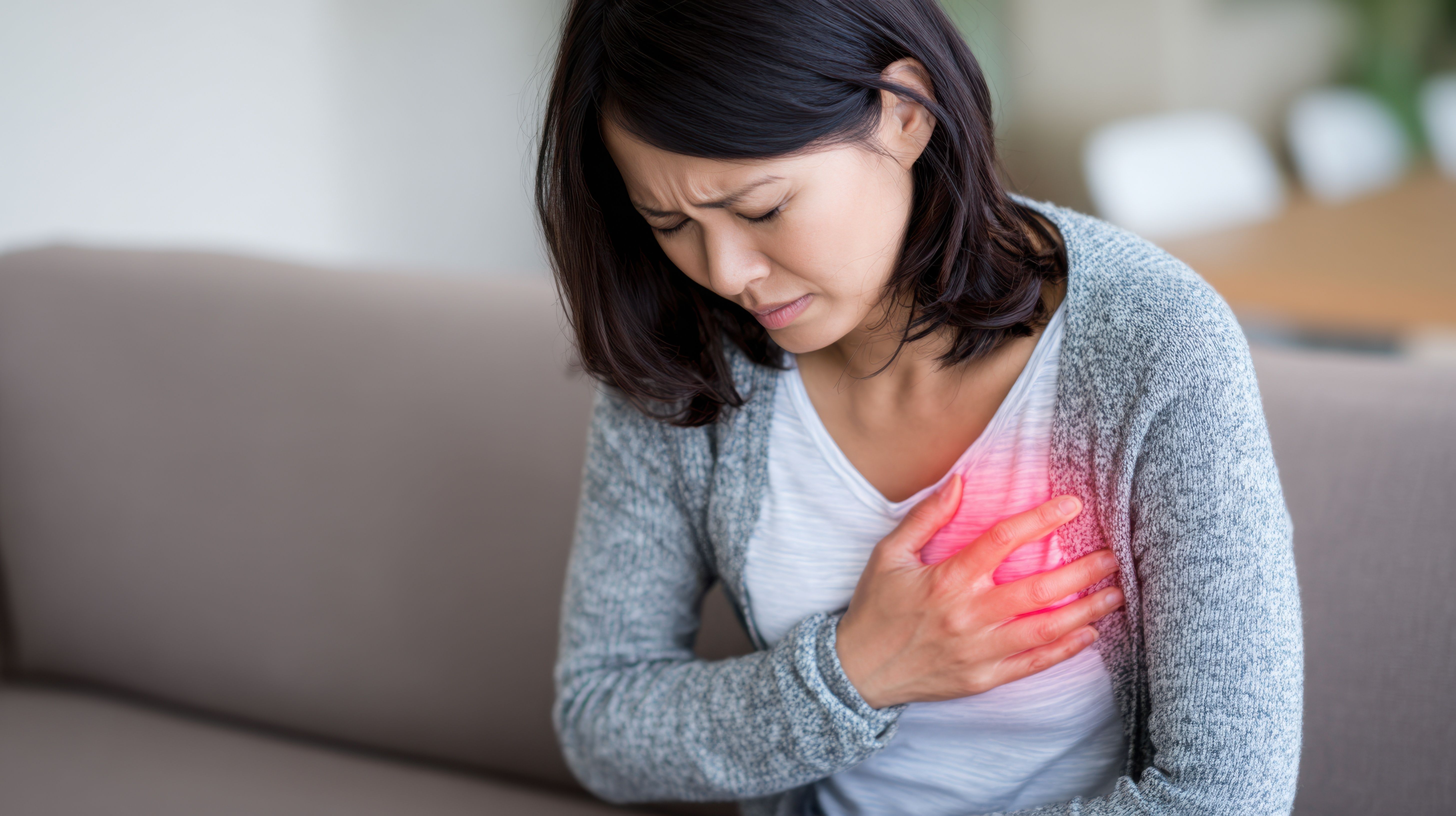 Woman holding chest in pain, experiencing discomfort, possible cardiac issue, sitting on a couch.