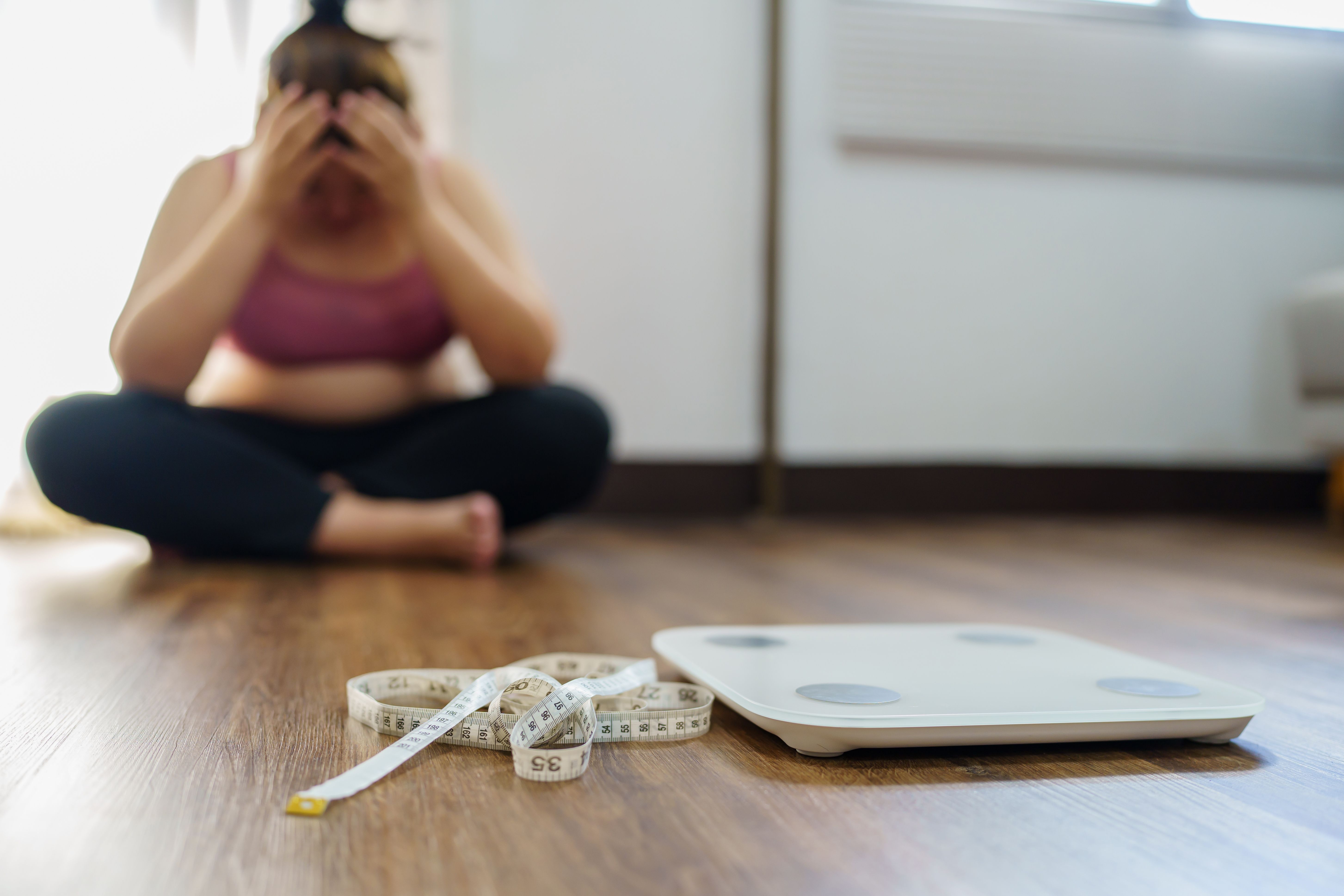 Obese Woman with fat upset bored of dieting Weight loss fail  Fat diet and scale sad asian woman on weight scale at home weight control - Image credit: Charlie's | stock.adobe.com