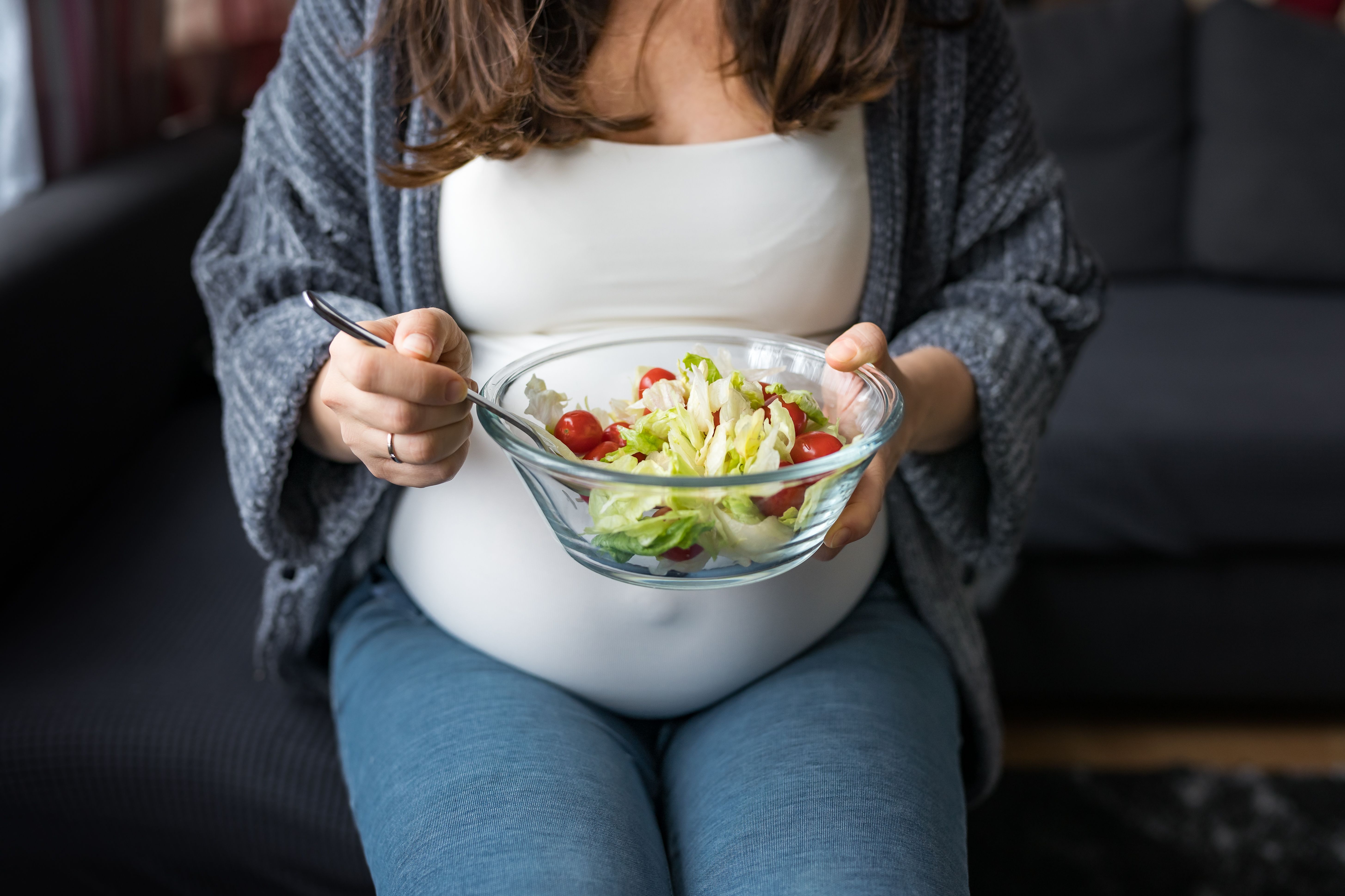 Pregnant woman eating fresh vegetable salad, sitting on a sofa, healthy pregnancy concept - Image credit: Anna | stock.adobe.com