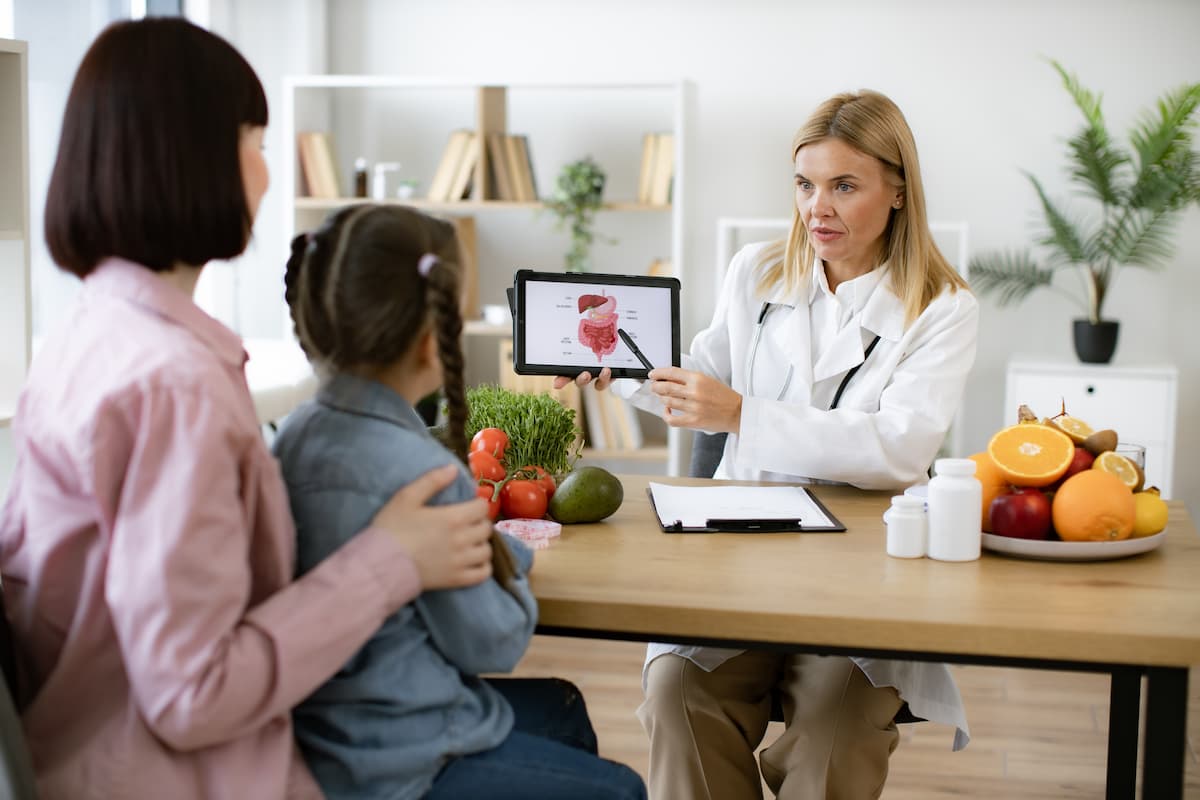 Mature experienced female doctor showing tablet with gastrointestinal tract image for treatment planning to little female child.