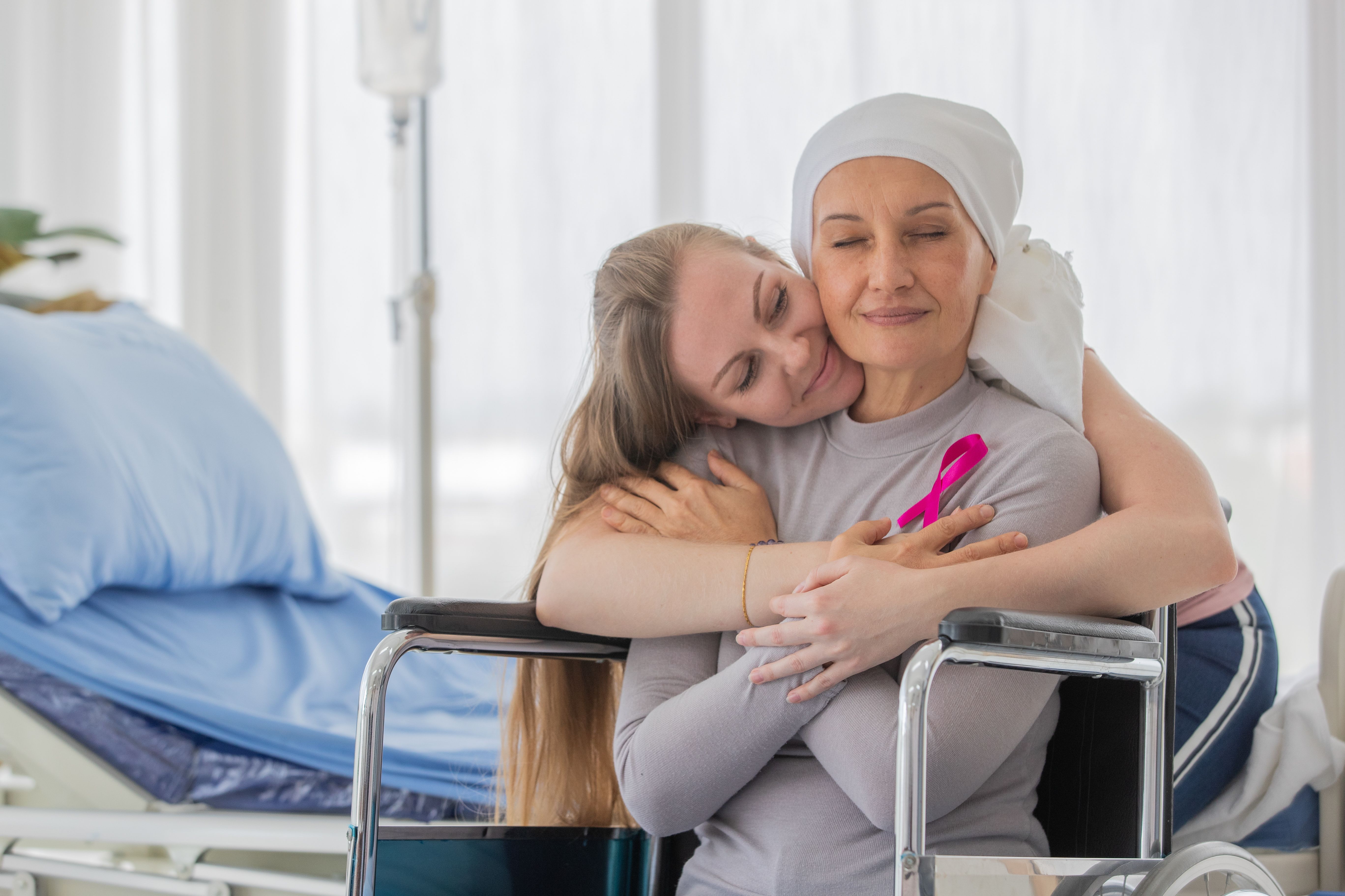 A middle-aged breast cancer woman with clothing around her head effected from chemo therapy sitting on wheel chair and hold hand of her daughter with hope and trust in love. | Image Credit: Bangkok Click Studio | stock.adobe.com