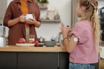 Back view of young girl with diabetes standing at kitchen counter in morning and chatting to mom focus on glucose monitoring device on arm copy space