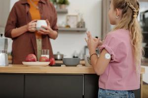 Back view of young girl with diabetes standing at kitchen counter in morning and chatting to mom focus on glucose monitoring device on arm copy space