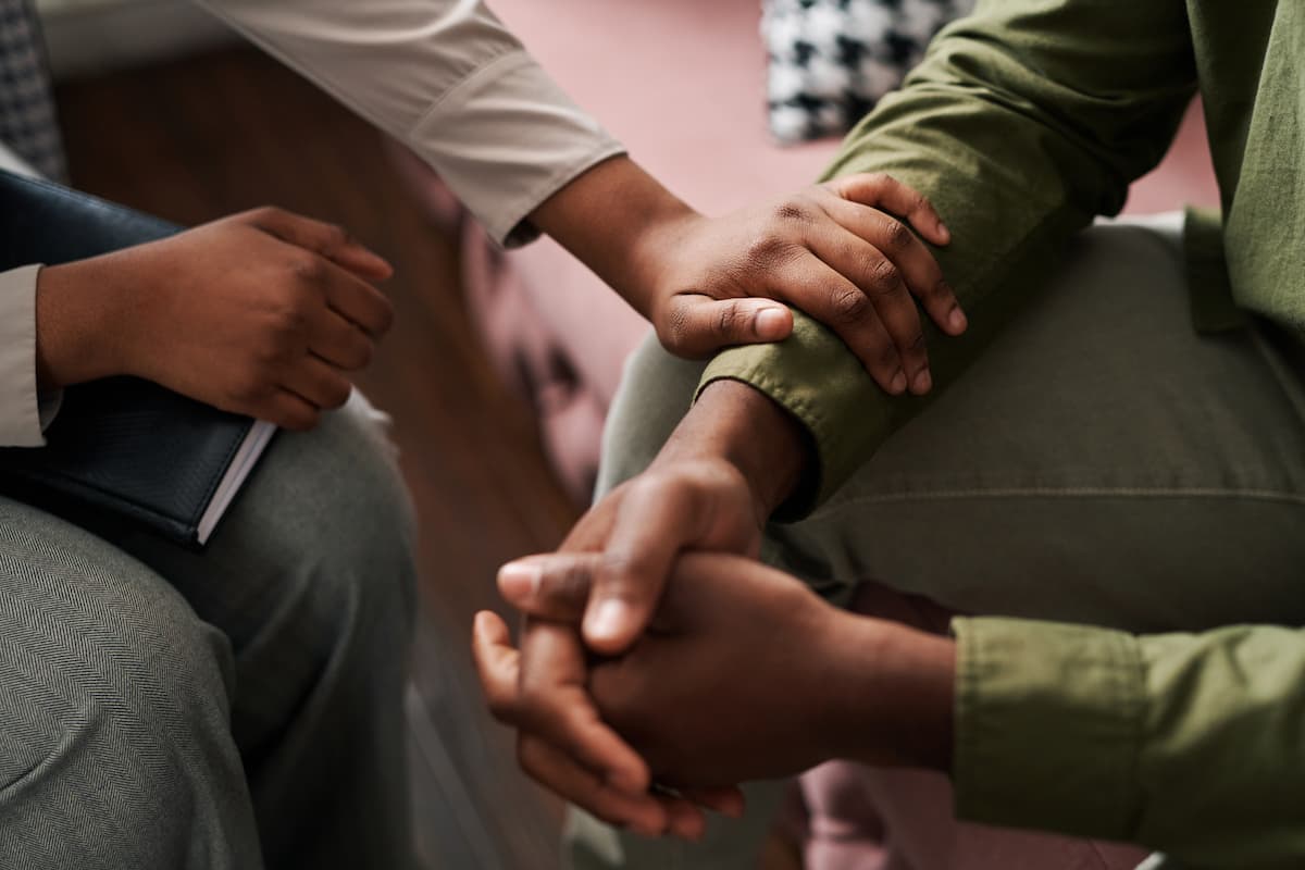 Young African American female psychologist keeping hand of wrist of male patient sitting in front of her and sharing his problems