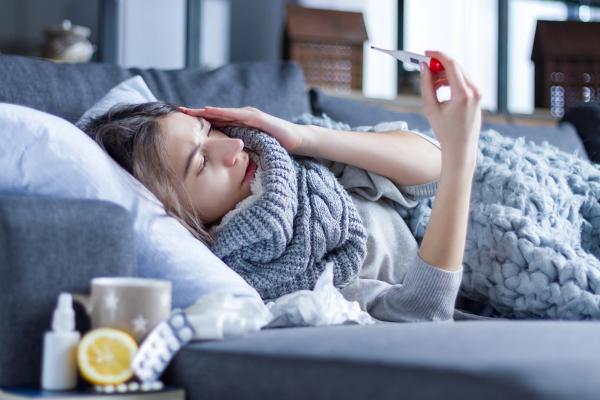 Sick exhausted girl in scarf is lying in bed wrapped in blanket. Young woman with fever and headache is measuring temperature with thermometer, treated at home. Winter cold and flu concept - Image credit: Monstar Studio | stock.adobe.com