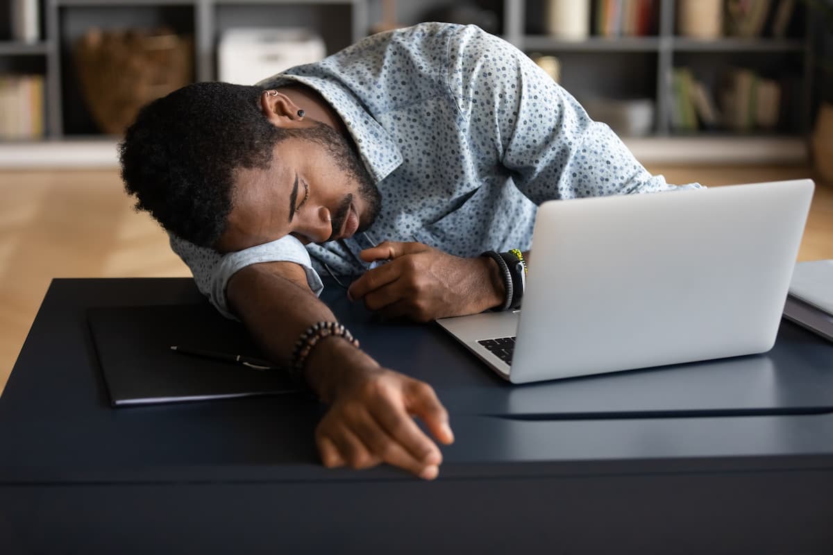 Exhausted tired African American businessman entrepreneur lying on desk with closed eyes, falling asleep, overworked young man unmotivated student sleeping at workplace, boring routine work