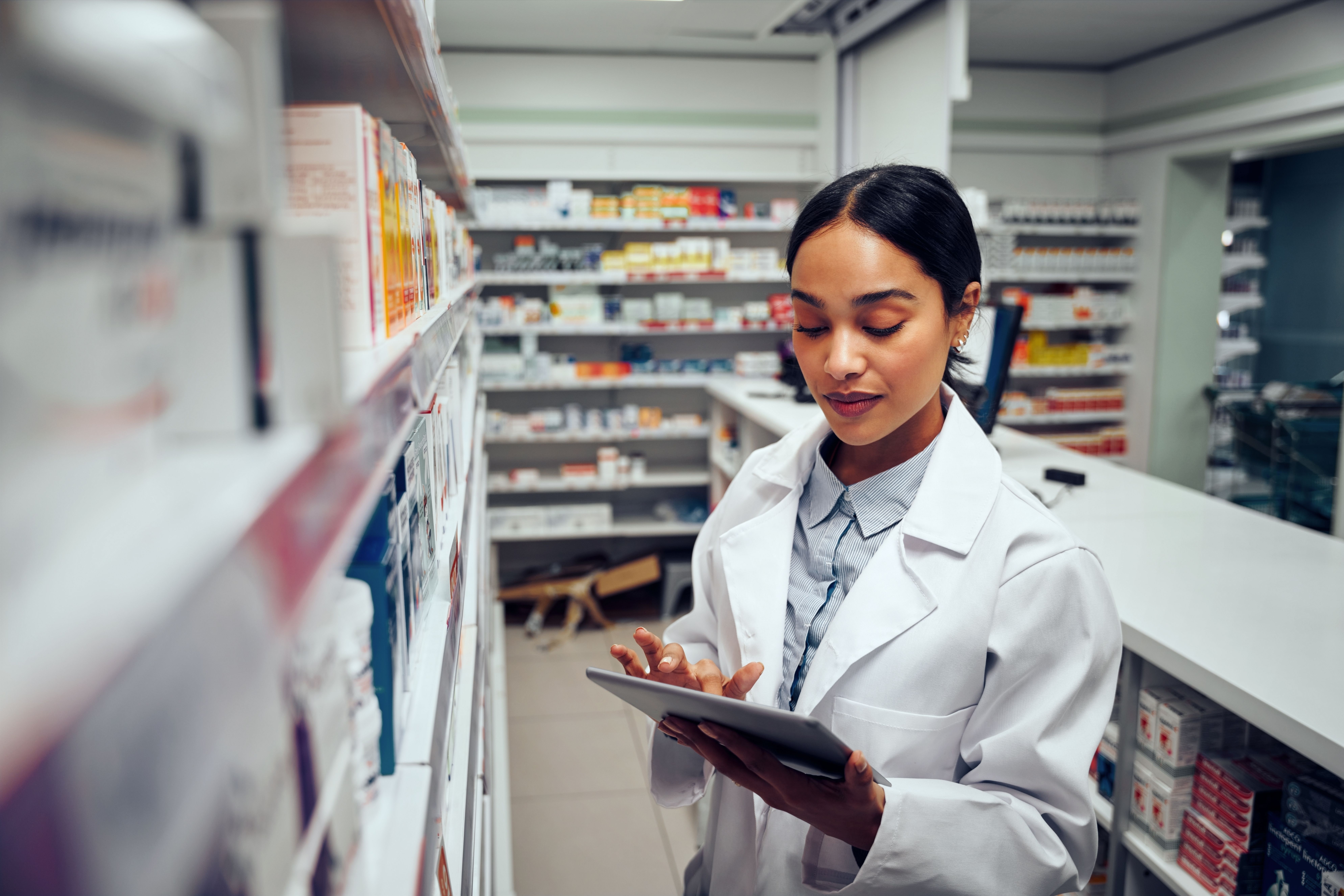 Young female pharmacist checking inventory of medicines in pharmacy using digital tablet wearing labcoat standing behind counter | Image Credit: StratfordProductions - stock.adobe.com