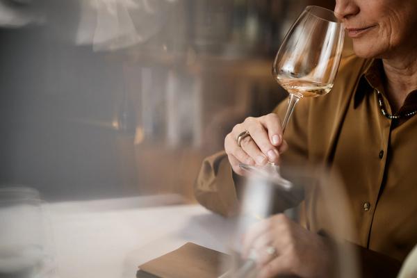 Middle aged Caucasian woman holding wine glass, gently swirling and smelling wine during tasting session, sitting at table with focus on hand and face, lips slightly smiling