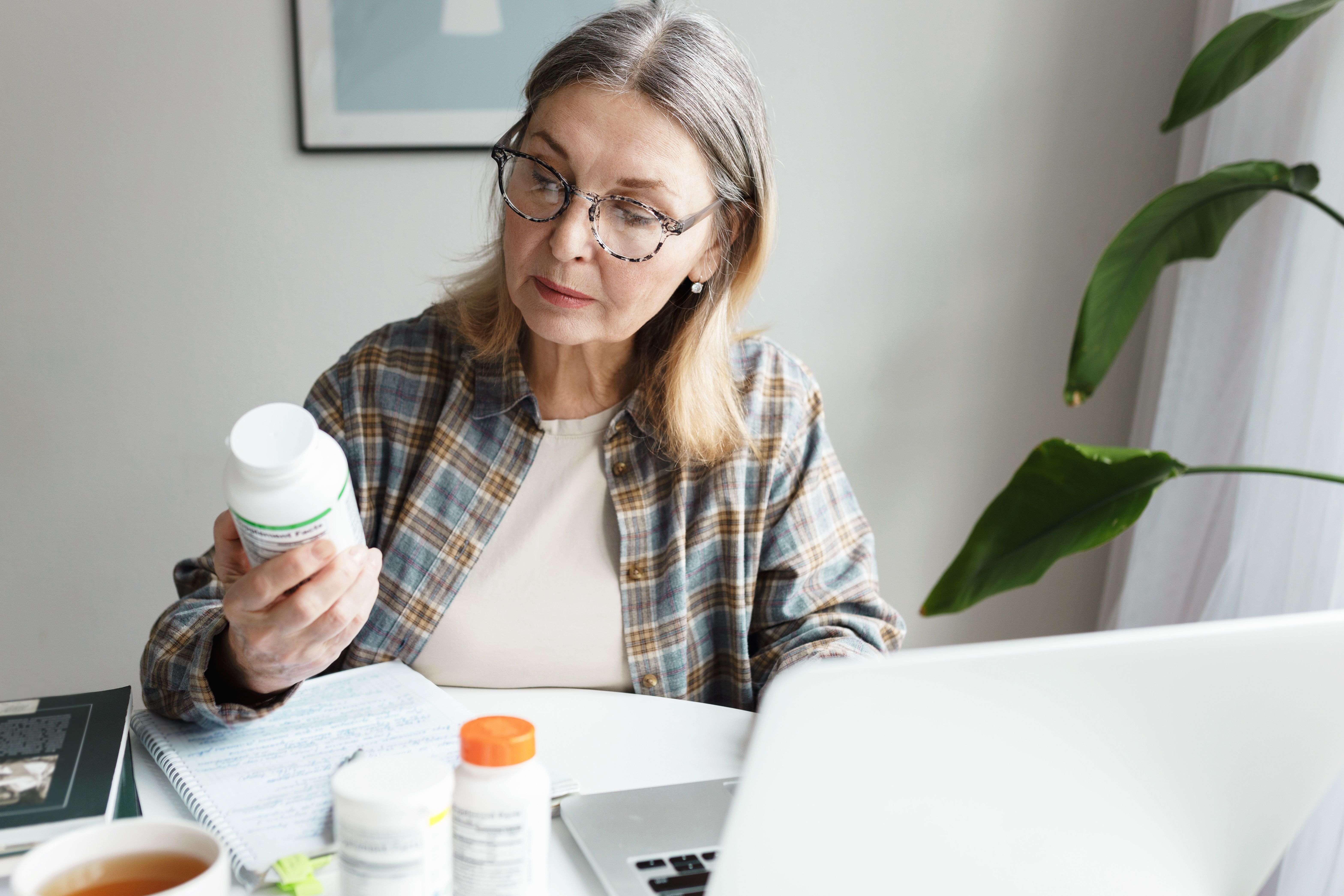 woman holding bottle of dietary supplement and reading label with instruction, ingredients or brand name carefully before ordering vitamins online using laptop and web application | Image Credit: shurkin_son | adobe.stock.com