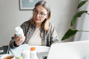 woman holding bottle of dietary supplement and reading label with instruction, ingredients or brand name carefully before ordering vitamins online using laptop and web application | Image Credit: shurkin_son | adobe.stock.com
