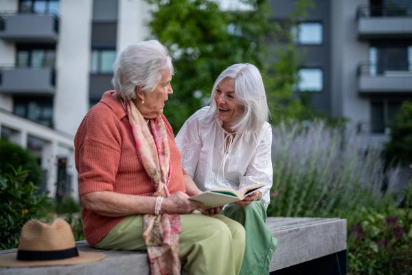 Elderly mother sitting on bench with adult daughter, reading together.