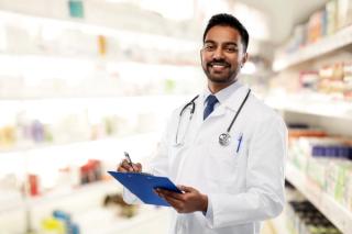 smiling indian male doctor or pharmacist in white coat with stethoscope and clipboard over drugstore background