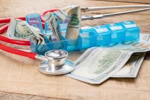 One-hundred-dollar bills stuffed into a blue pill organizer, representing the high cost of medicine in the United States. | Image Credit: Deborah | adobe.stock.com