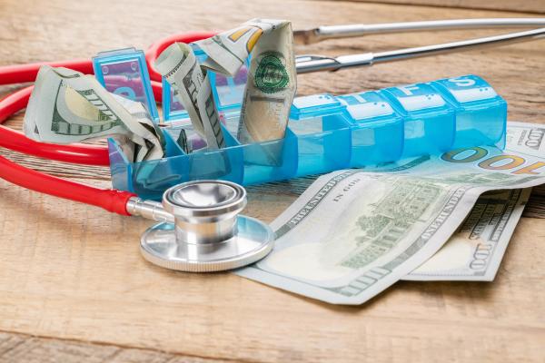 One-hundred-dollar bills stuffed into a blue pill organizer, representing the high cost of medicine in the United States. | Image Credit: Deborah | adobe.stock.com