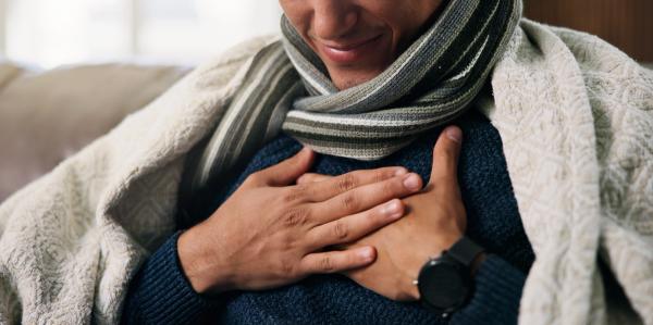 Hands, chest pain and sick on sofa with blanket for flu, rest and recovery in lounge at house. Person, respiratory illness and cold symptoms with congestion, comfort or healing in living room at home