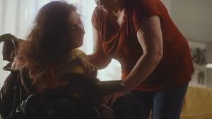 Aged woman caregiver helping woman with a spinal muscular atrophy setting a modern motorized wheelchair for comfortable sitting