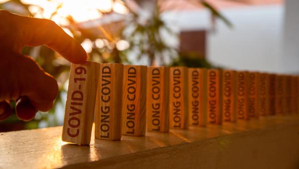 Hand poked on a row of wooden dominoes, with the words "COVID19" on the first piece and the words "LONG COVID" on subsequent pieces - Image credit: Anucha | stock.adobe.com