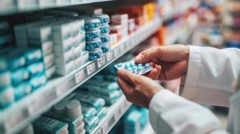 Hands of a pharmacist or healthcare professional holding a blister pack of capsules in front of a pharmacy shelf stocked with various medications - Image credit: MP Studio | stock.adobe.com