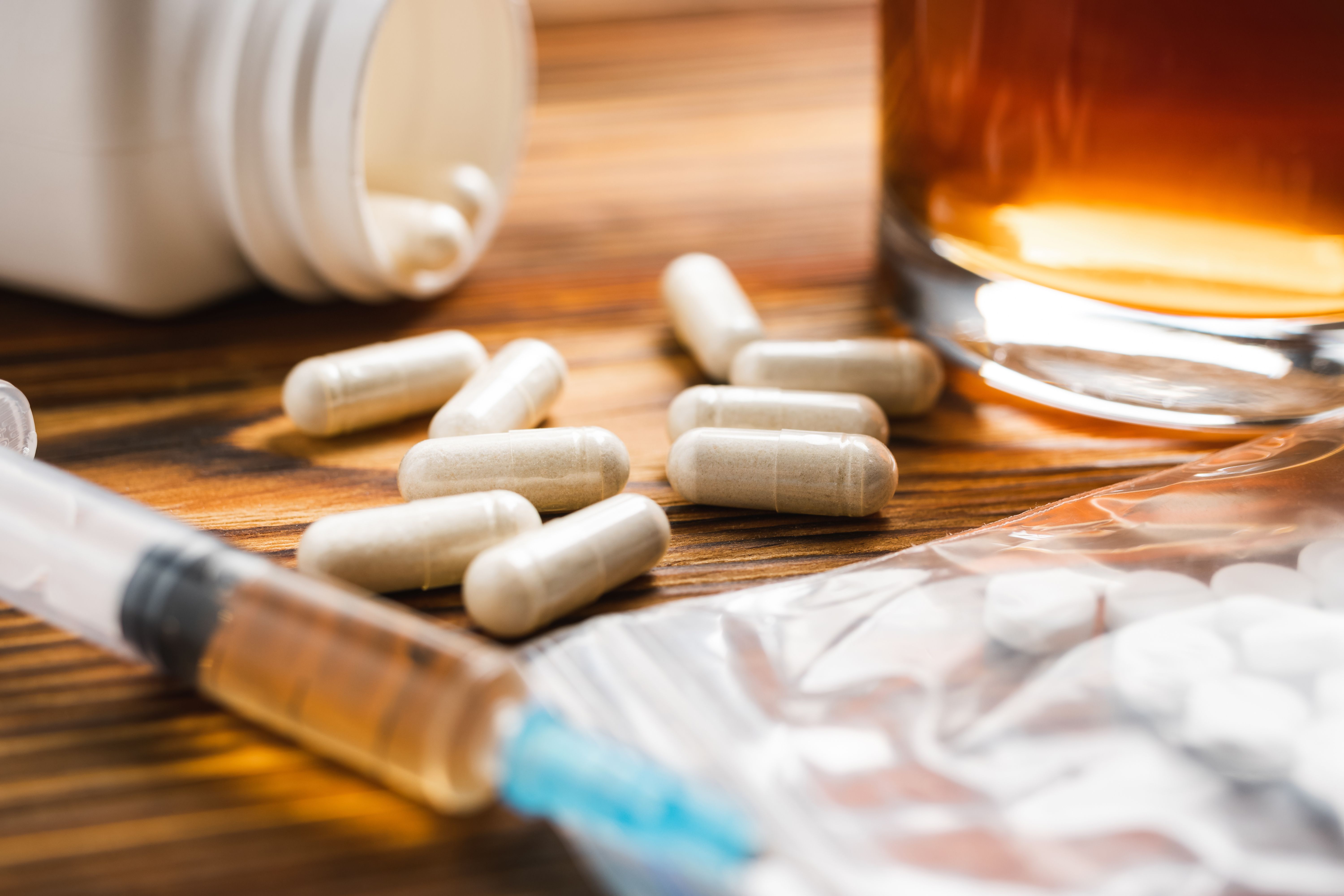 White pills, syringe with a drugs dose, narcotics, glass with alcohol drink on a wooden table, close-up view. Concept of addiction and bad habits - Image credit: O.Farion | stock.adobe.com