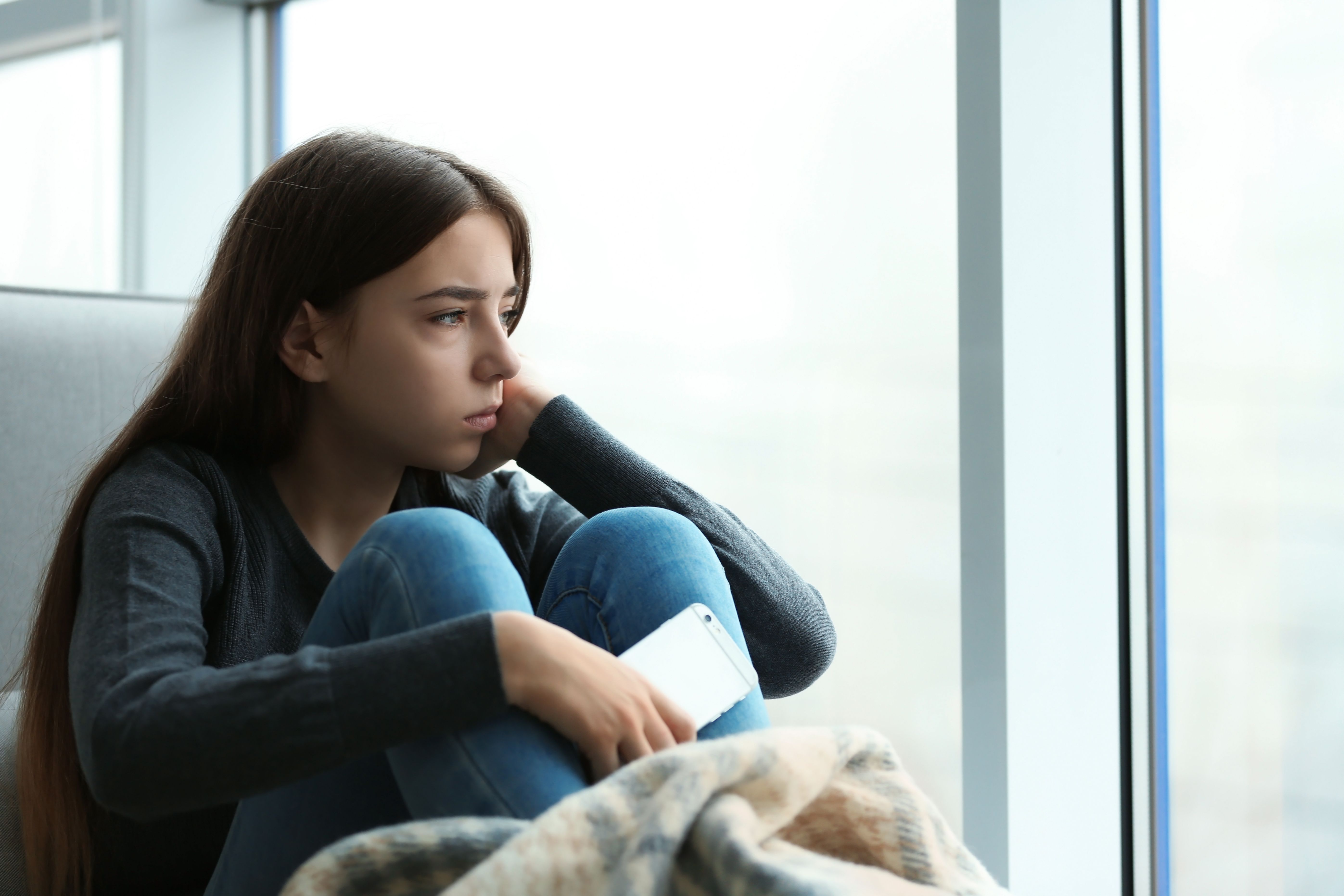 Upset teenage girl with smartphone sitting at window indoors - Image credit: New Africa | stock.adobe.com