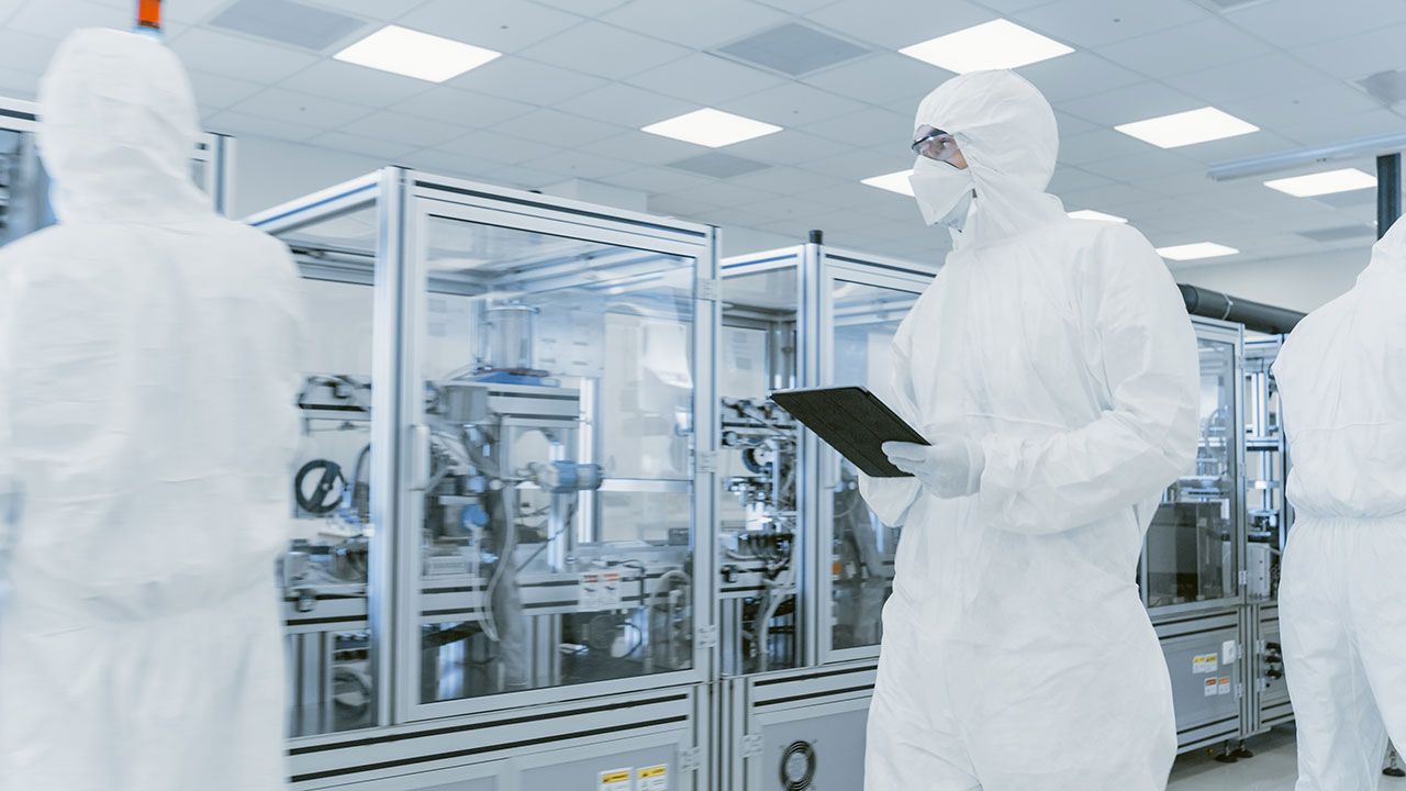 Quality Control Check: Scientist Using Digital Tablet Computer and wearing Protective Suit walks through Manufacturing Laboratory. Product Manufacturing: Pharmaceutics, Semiconductors | Image Credit: ©Gorodenkoff -stock.adobe.com