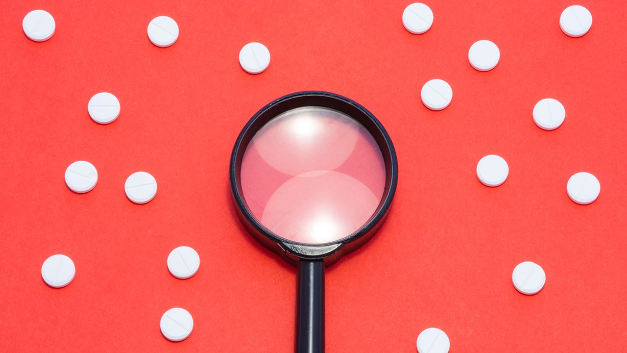 Black magnifying glass is on red background surrounded by white pills as ornament polka dots. Medical or pharmaceutical concept by pharmacological drugs quality control, identification of drugs,pills| Image Credit: © shidlovski – Stock.adobe.com