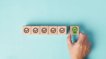 A hand is placing the final wooden block with a green certificate checkmark, symbolizing achievement, quality assurance and completion, against a plain pastel blue background. | Image credit: TimmyTimTim – stock.adobe.com