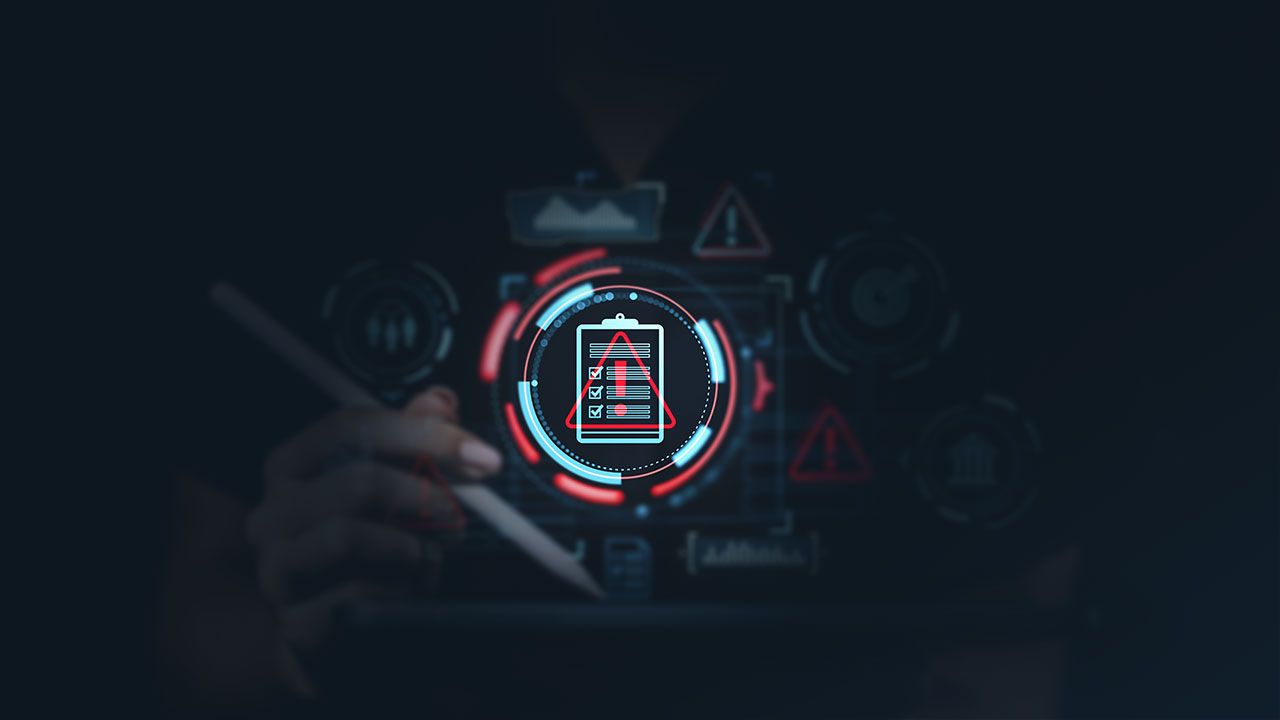 Hand holding a stylus over a tablet with futuristic risk management icons such as warning signs, data charts, and safety symbols on a dark background. | Image Credit: ©ImageFlow -stock.adobe.com