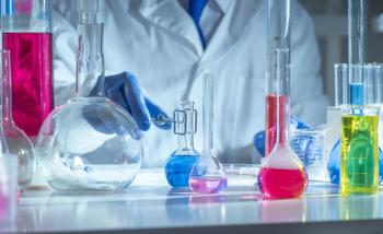 Chemistry laboratory scene with colorful chemical solutions in glass flasks and cylinders, scientist in gloves writing results, analytical experiment workspace. | Image Credit: © TSViPhoto - stock.adobe.com