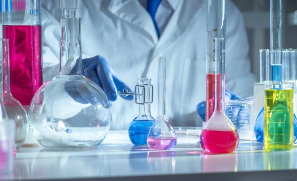 Chemistry laboratory scene with colorful chemical solutions in glass flasks and cylinders, scientist in gloves writing results, analytical experiment workspace. | Image Credit: © TSViPhoto - stock.adobe.com